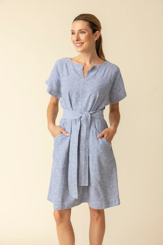 A woman wearing the Habitat Bethany Dress with Belt in light blue, featuring short sleeves, knee-length hem, tie belt, and pockets, stands against a plain beige background.