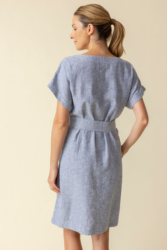 A woman wearing the Habitat Bethany Dress with Belt in light blue, featuring short sleeves, knee-length hem, tie belt, and pockets, stands against a plain beige background.