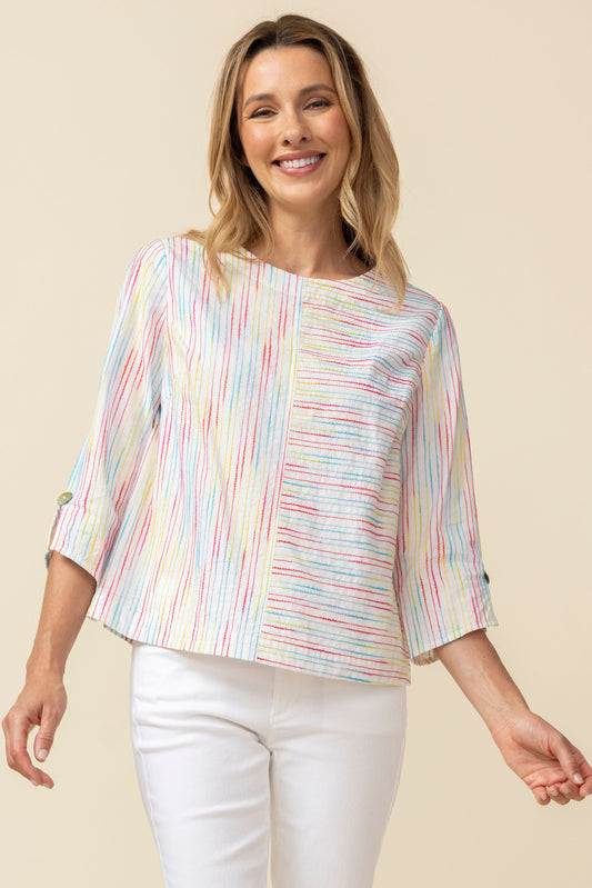 A woman smiles while wearing the Habitat Carousel Pullover, featuring a multi-color design and 3/4 sleeves, paired with white pants against a plain beige background.