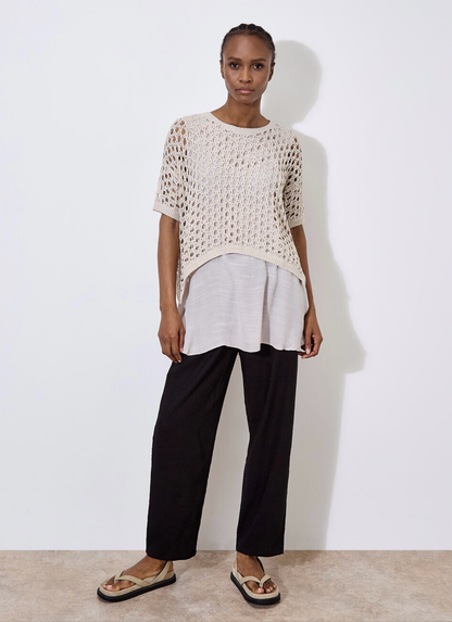 Against a plain, light backdrop, the individual wears Apricot's Knit Mesh Slub Tunic over a white blouse. This textured cream-crochet top features short sleeves and adds elegance as they gaze toward the camera.