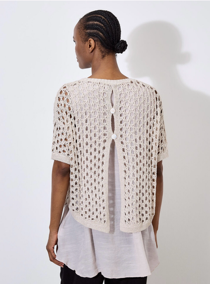 Against a plain, light backdrop, the individual wears Apricot's Knit Mesh Slub Tunic over a white blouse. This textured cream-crochet top features short sleeves and adds elegance as they gaze toward the camera.