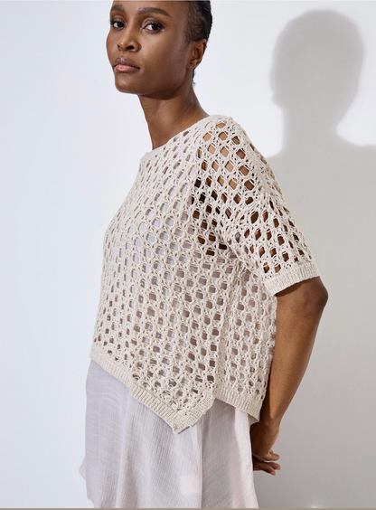 Against a plain, light backdrop, the individual wears Apricot's Knit Mesh Slub Tunic over a white blouse. This textured cream-crochet top features short sleeves and adds elegance as they gaze toward the camera.
