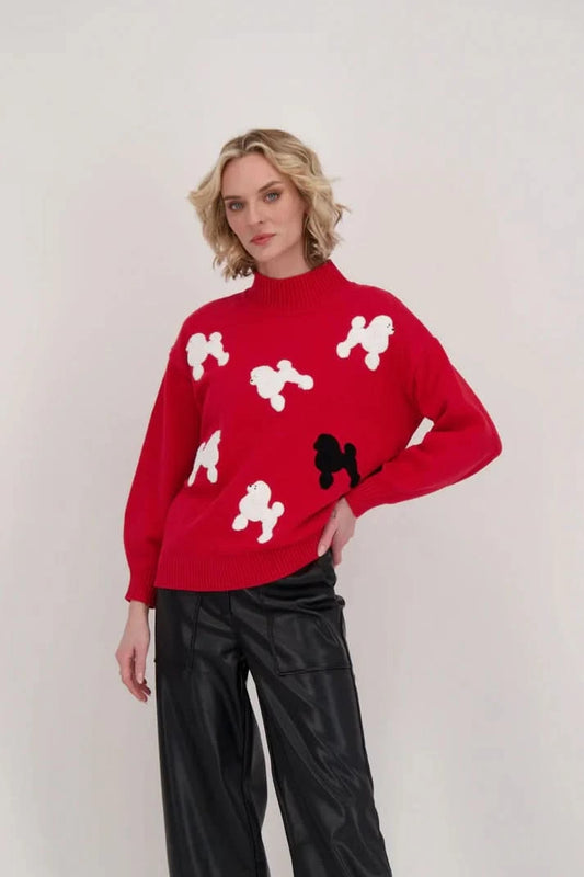 Against a plain backdrop, a poodle enthusiast exhibits their fashion sense by wearing the Poodle Turtleneck Sweater With Back-Tie Detail from Gabby Isabella—a striking red sweater embellished with white and black poodle motifs—and pairs it seamlessly with black pants.