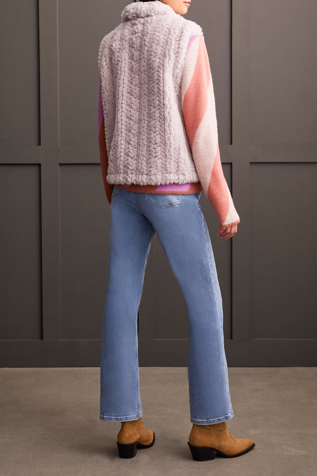 A person wearing a pink and white sweater, a Tribal Fluffy Sleeveless Vest in light gray, light blue jeans, and brown heeled boots stands indoors against a paneled wall, embodying fashion with comfort.