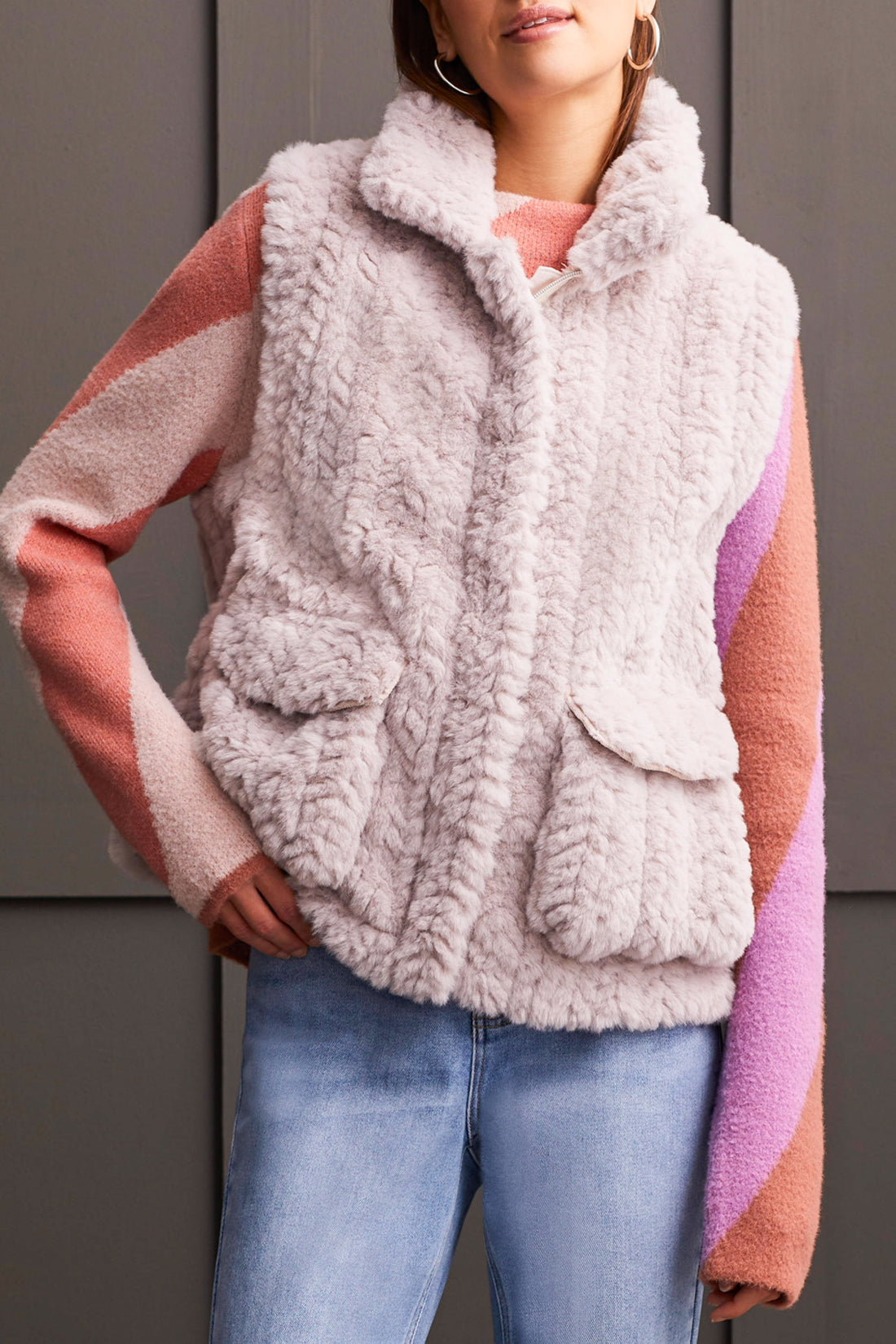 A person wearing a pink and white sweater, a Tribal Fluffy Sleeveless Vest in light gray, light blue jeans, and brown heeled boots stands indoors against a paneled wall, embodying fashion with comfort.