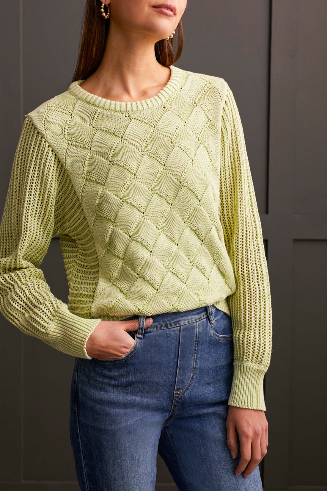 A person is wearing a light green Dolman Crew Neck Sweater with Special Wash from Tribal, along with blue jeans, standing against a dark wall. The luxurious comfort of the soft fabric is evident as the person's hand rests casually in one pocket.