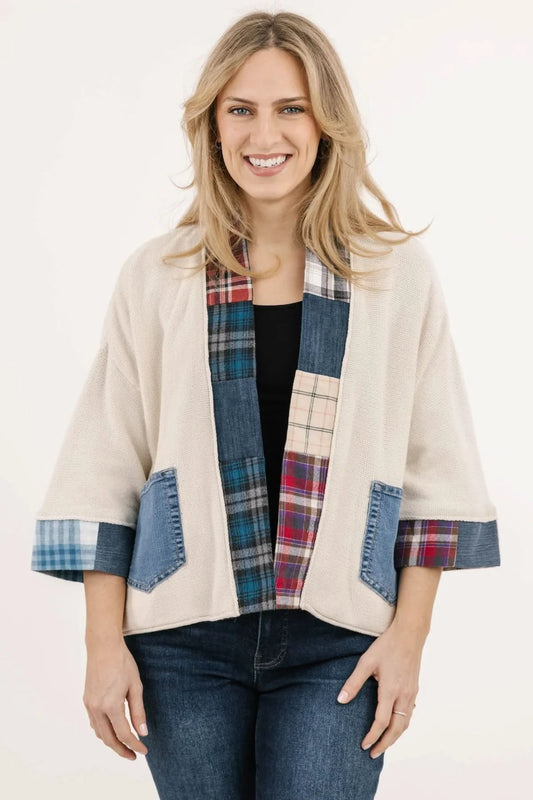 A woman stands sideways against a stone wall wearing the Alpine Love Cardigan by Shannon Passero—an upcycled flannel with a plaid heart patch on the back, plaid sleeve details, and blue jeans for a chic, sustainable look.