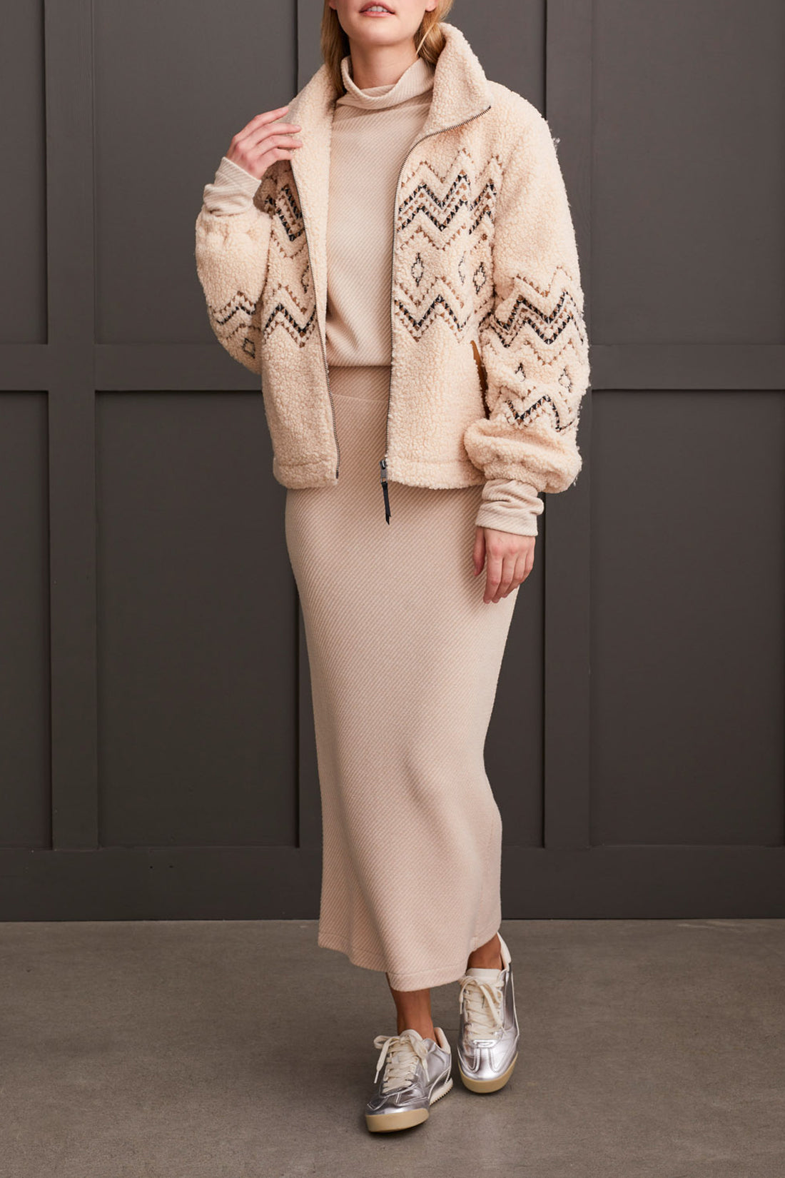 A person wearing a Tribal Lined Zip up Jacket with Embroidery in beige, paired with a matching ribbed dress, standing against a dark background.