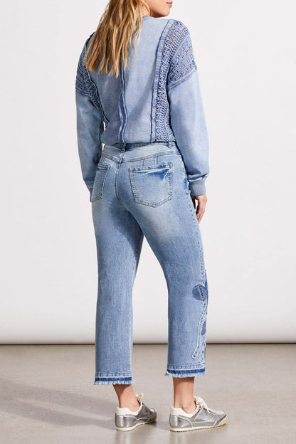 A person is wearing a light blue, textured sweater, Tribal's Sophia Microflare Crop with Denim jeans featuring embroidery near the hem, and silver sneakers. The image captures the outfit from head to lower legs.