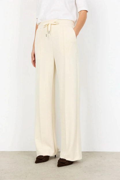 A person wearing Soya Concepts Banu Pant in cream, featuring an adjustable elastic drawstring waist, paired with a white top and brown pointed-toe shoes, stands against a plain background.