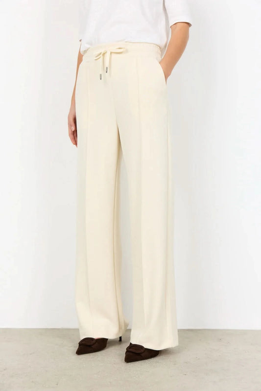 A person wearing Soya Concepts Banu Pant in cream, featuring an adjustable elastic drawstring waist, paired with a white top and brown pointed-toe shoes, stands against a plain background.