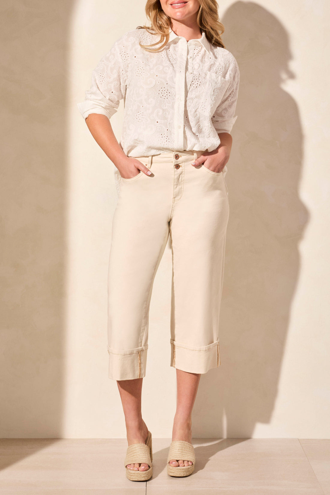 A woman stands against a neutral wall wearing a white eyelet blouse and woven sandals, paired with Tribal's BROOKE GIRLFRIEND WIDE JEANS WITH CUFFS.