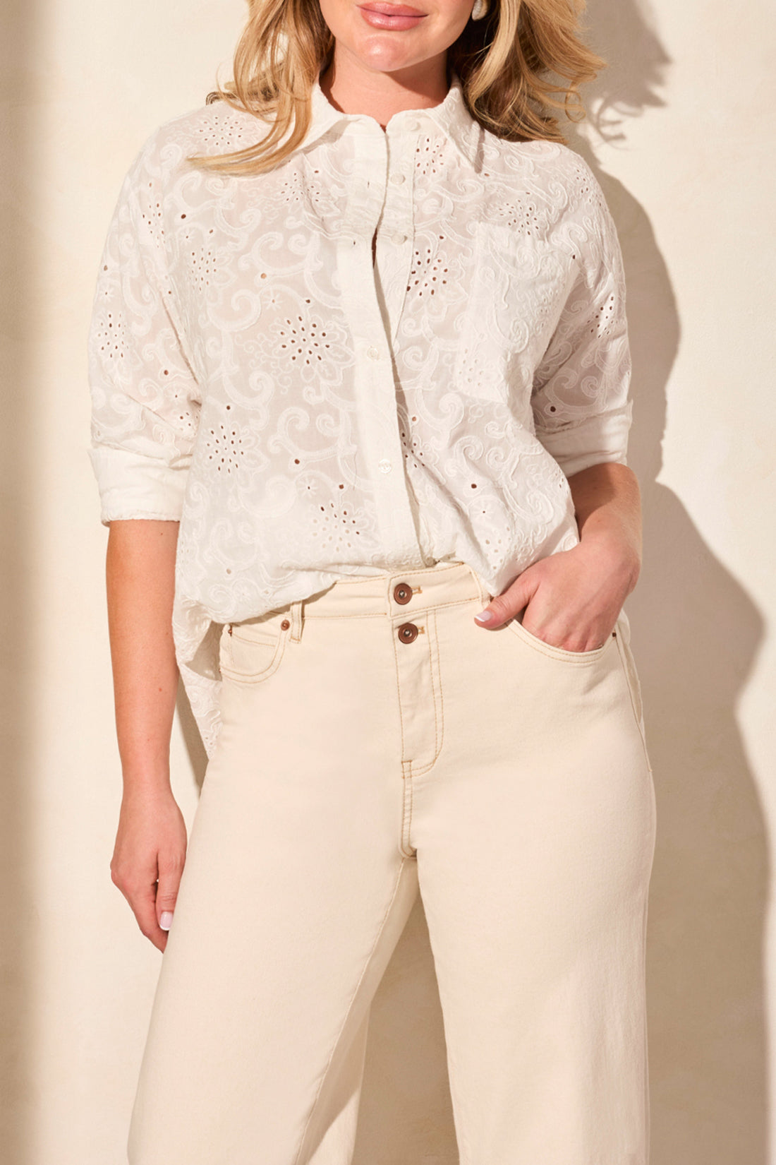 A woman stands against a neutral wall wearing a white eyelet blouse and woven sandals, paired with Tribal's BROOKE GIRLFRIEND WIDE JEANS WITH CUFFS.