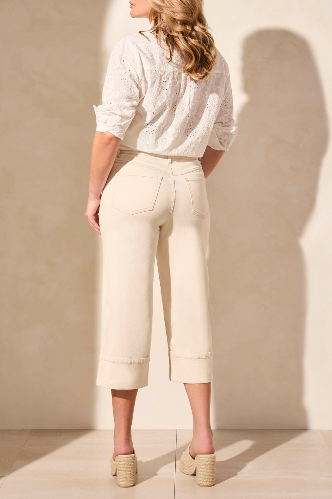 A woman stands against a neutral wall wearing a white eyelet blouse and woven sandals, paired with Tribal's BROOKE GIRLFRIEND WIDE JEANS WITH CUFFS.