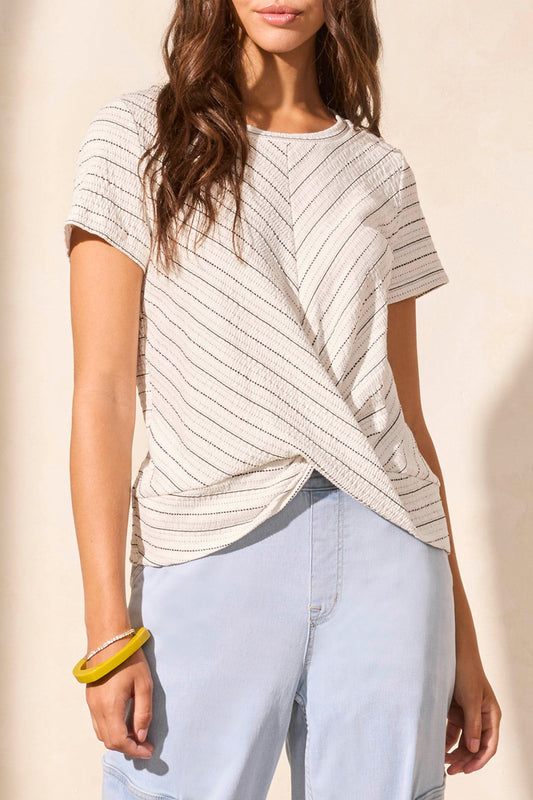 A woman models the Tribal SHORT SLEEVE CREW NECK TWIST TOP paired with light blue pants and a yellow bracelet against a beige backdrop, embodying a stylish wardrobe upgrade.