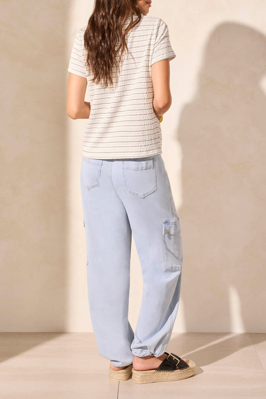 A woman models the Tribal SHORT SLEEVE CREW NECK TWIST TOP paired with light blue pants and a yellow bracelet against a beige backdrop, embodying a stylish wardrobe upgrade.