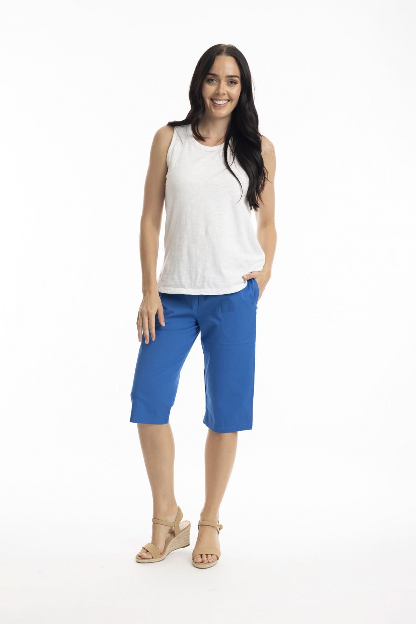 A woman stands smiling in a white sleeveless top, beige sandals, and Orientique Bangalene Shorts With Pockets—blue, knee-length shorts featuring a flexible fit—against a plain white background.