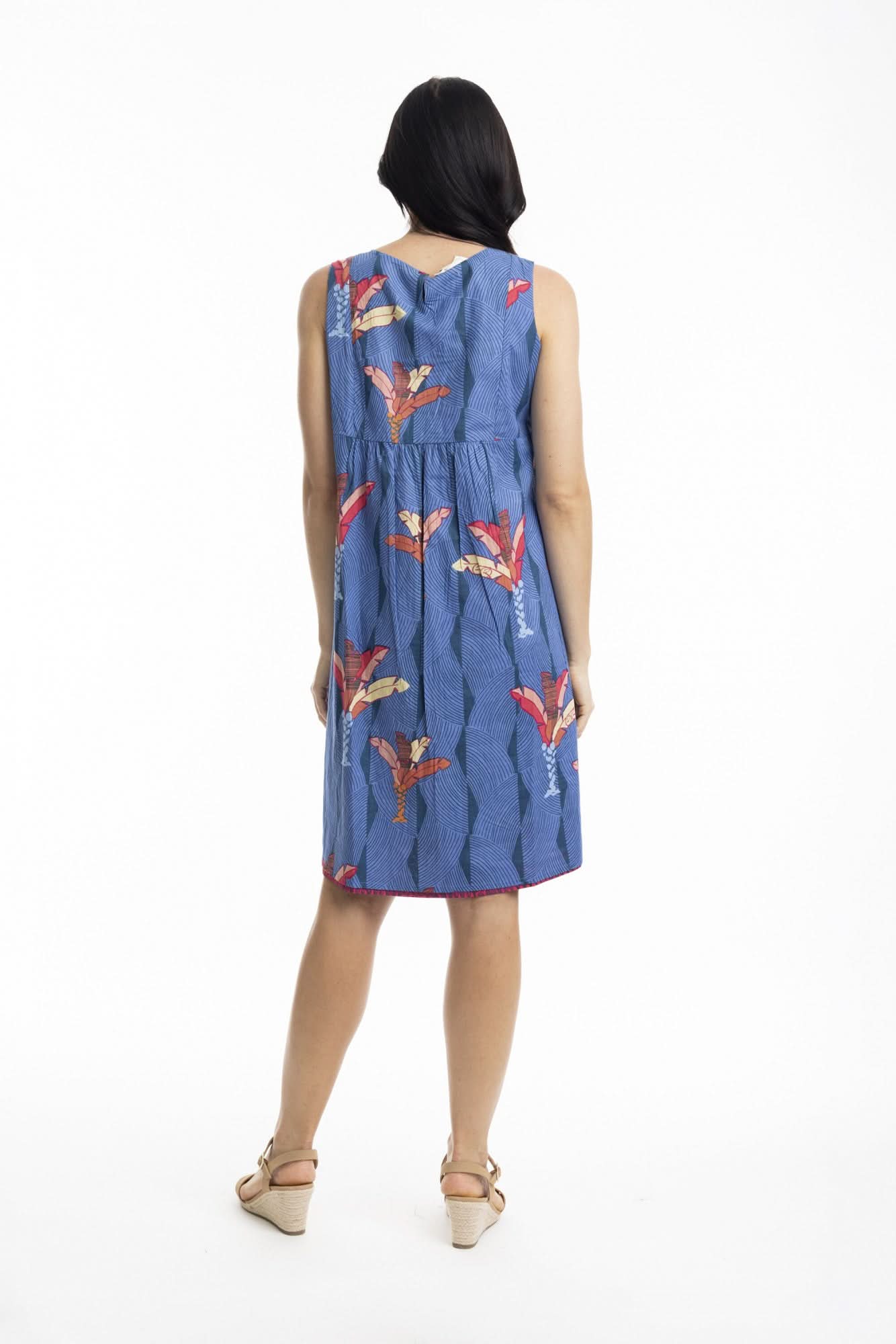 A woman stands in profile wearing the Orientique El Gouna Reversible Shift Dress, a blue dress with red and orange floral patterns and pockets, styled with beige wedge sandals against a plain white background.