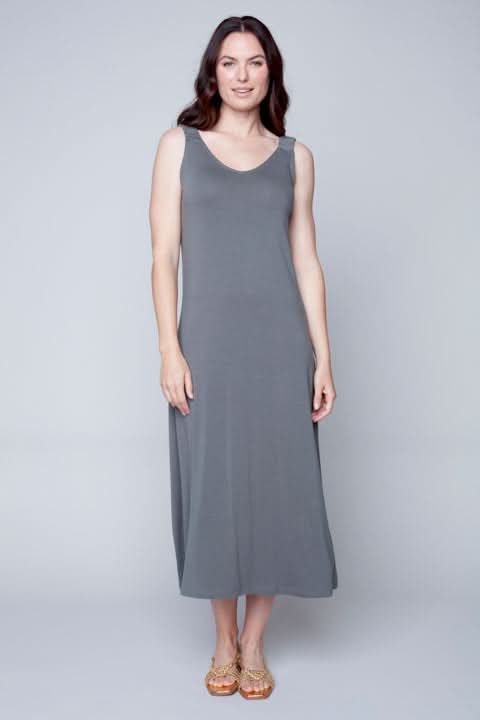A woman standing against a plain background wearing a Sleeveless Flow Dress by Carre Noir made of soft fabric and embellished sandals.