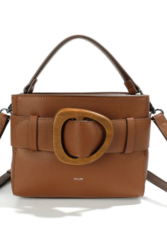 The Woodland Riley by Co-Lab is a brown leather handbag with a top handle, wide strap featuring a large wooden buckle, and a detachable shoulder strap.