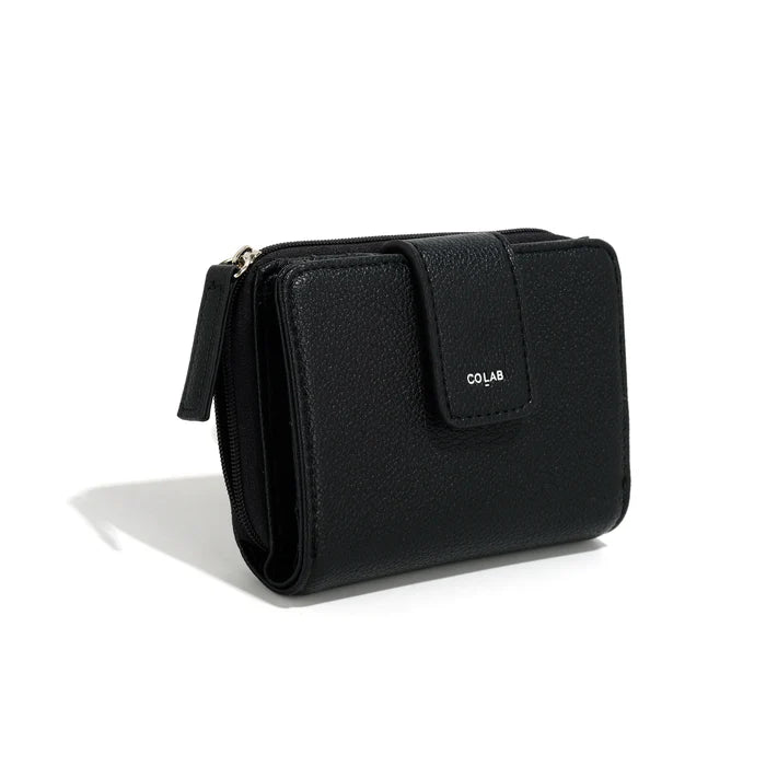 A person in a light gray shirt and beige pants holds The Lucie Small Wallet by Co-Lab, an eco-friendly, compact green wallet with both zipper and snap closures.