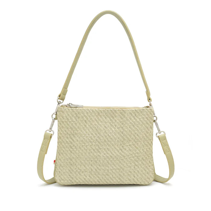 A woman stands against a plain background wearing a light gray blouse, beige pants, and The Sandrine Shoulder / Crossbody by Co-Lab—crafted from recycled vegan leather with an adjustable strap.