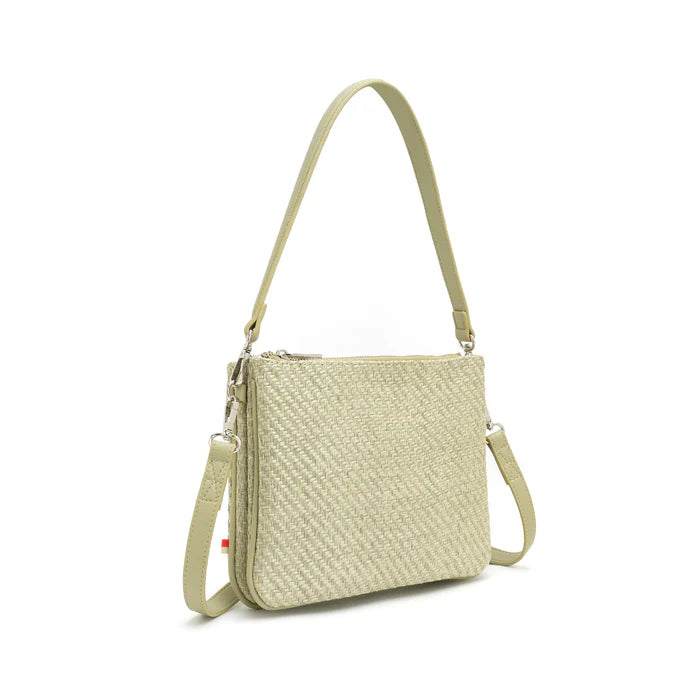 A woman stands against a plain background wearing a light gray blouse, beige pants, and The Sandrine Shoulder / Crossbody by Co-Lab—crafted from recycled vegan leather with an adjustable strap.