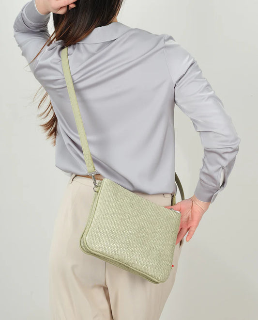 A woman stands against a plain background wearing a light gray blouse, beige pants, and The Sandrine Shoulder / Crossbody by Co-Lab—crafted from recycled vegan leather with an adjustable strap.