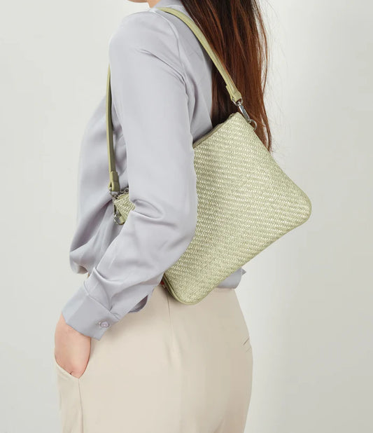A woman stands against a plain background wearing a light gray blouse, beige pants, and The Sandrine Shoulder / Crossbody by Co-Lab—crafted from recycled vegan leather with an adjustable strap.