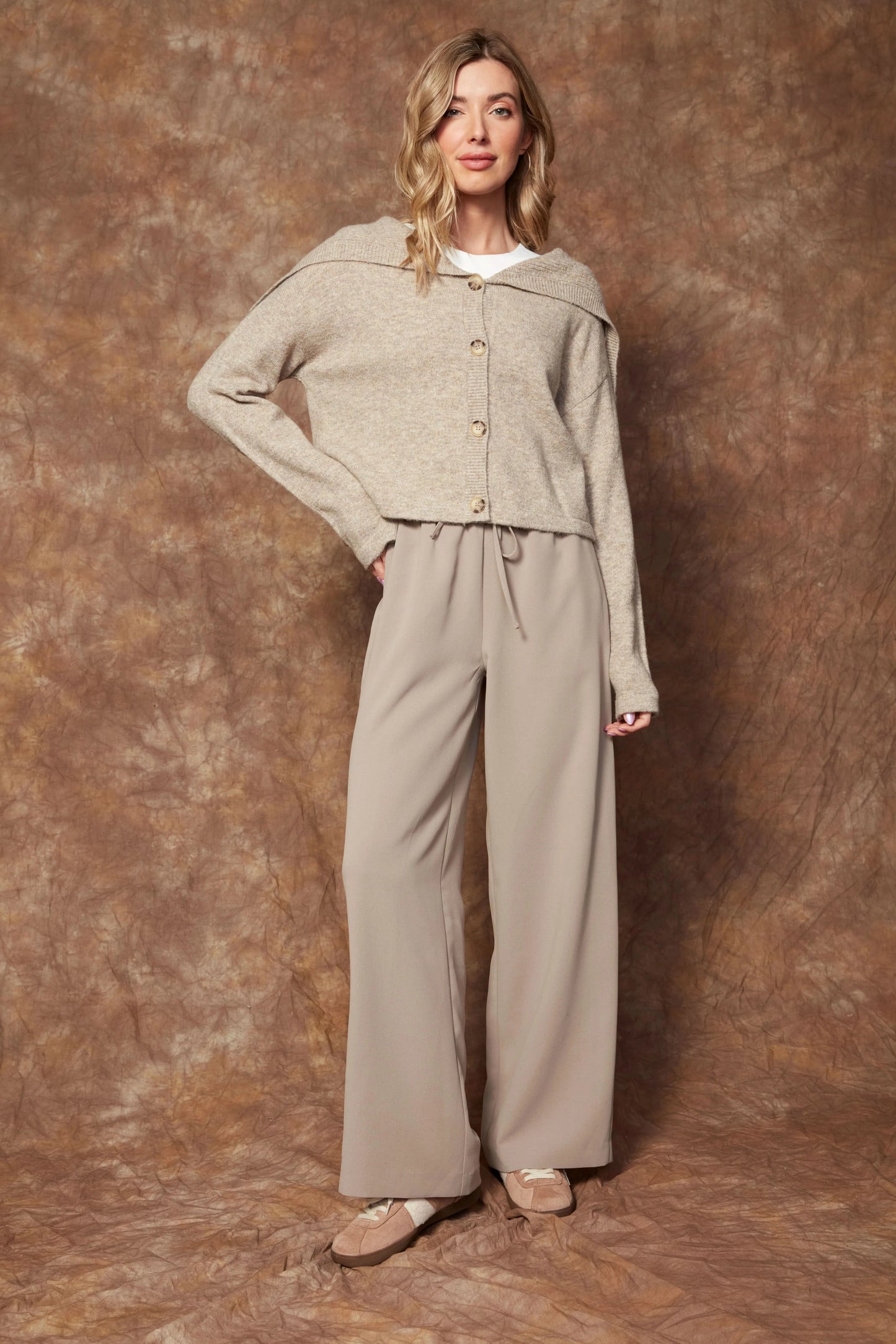 A woman stands before a brown textured backdrop, wearing RD International's Pull on Drawstring Pant in beige, paired with a beige cardigan, white shirt, and beige ballet flats.