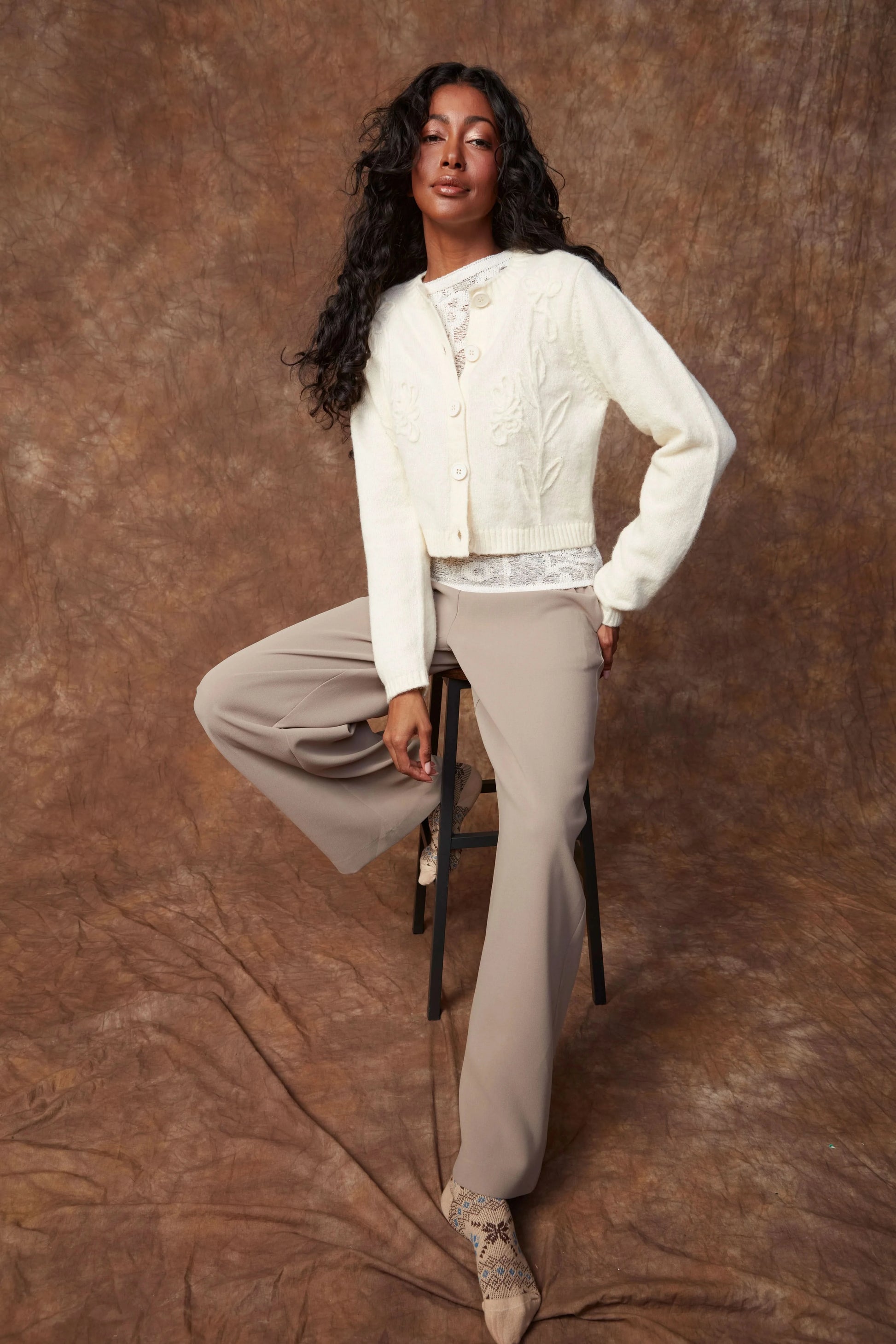 A woman stands before a brown textured backdrop, wearing RD International's Pull on Drawstring Pant in beige, paired with a beige cardigan, white shirt, and beige ballet flats.