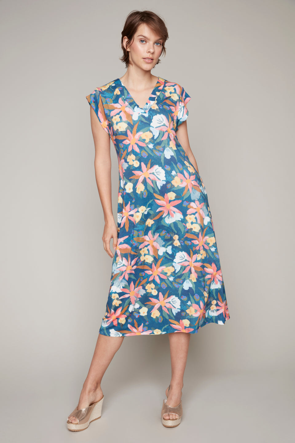 A person stands against a plain background wearing the Carre Noir Blue Floral V Neck Dress, styled with beige wedge sandals.