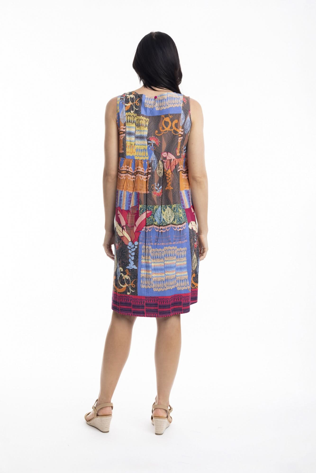 A woman stands in profile wearing the Orientique El Gouna Reversible Shift Dress, a blue dress with red and orange floral patterns and pockets, styled with beige wedge sandals against a plain white background.