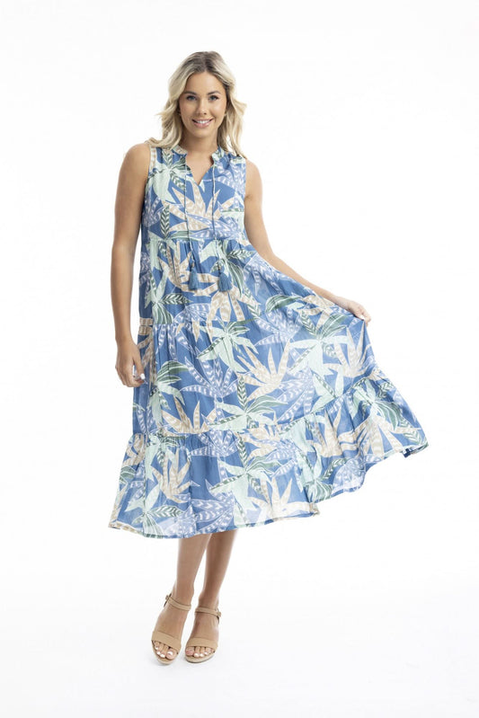 A woman poses against a white backdrop in the Orientique Fitzroy Sleeveless Boho Dress, featuring a blue tropical leaf print and v-neckline, paired with tan wedges—an ideal choice for summer events.