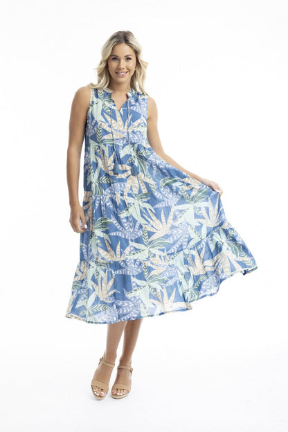 A woman poses against a white backdrop in the Orientique Fitzroy Sleeveless Boho Dress, featuring a blue tropical leaf print and v-neckline, paired with tan wedges—an ideal choice for summer events.