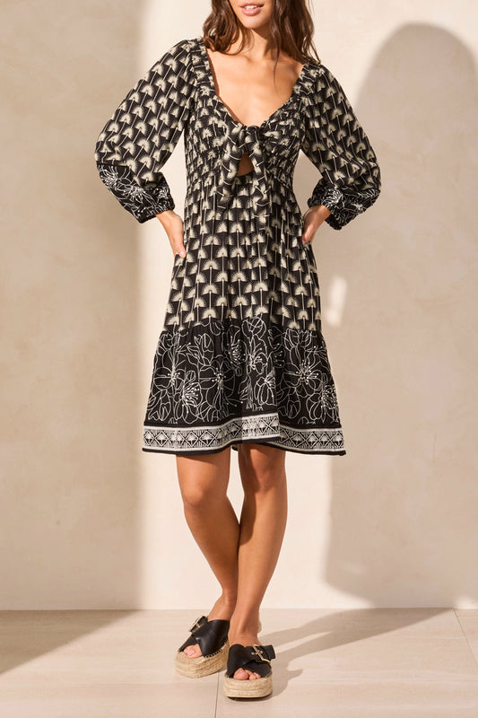 The woman confidently stands with hands on hips in a Tribal WEAR 2 WAYS EMBROIDERED DRESS, a versatile black and white patterned knee-length piece featuring long sleeves and a tie-front detail, paired perfectly with platform sandals.