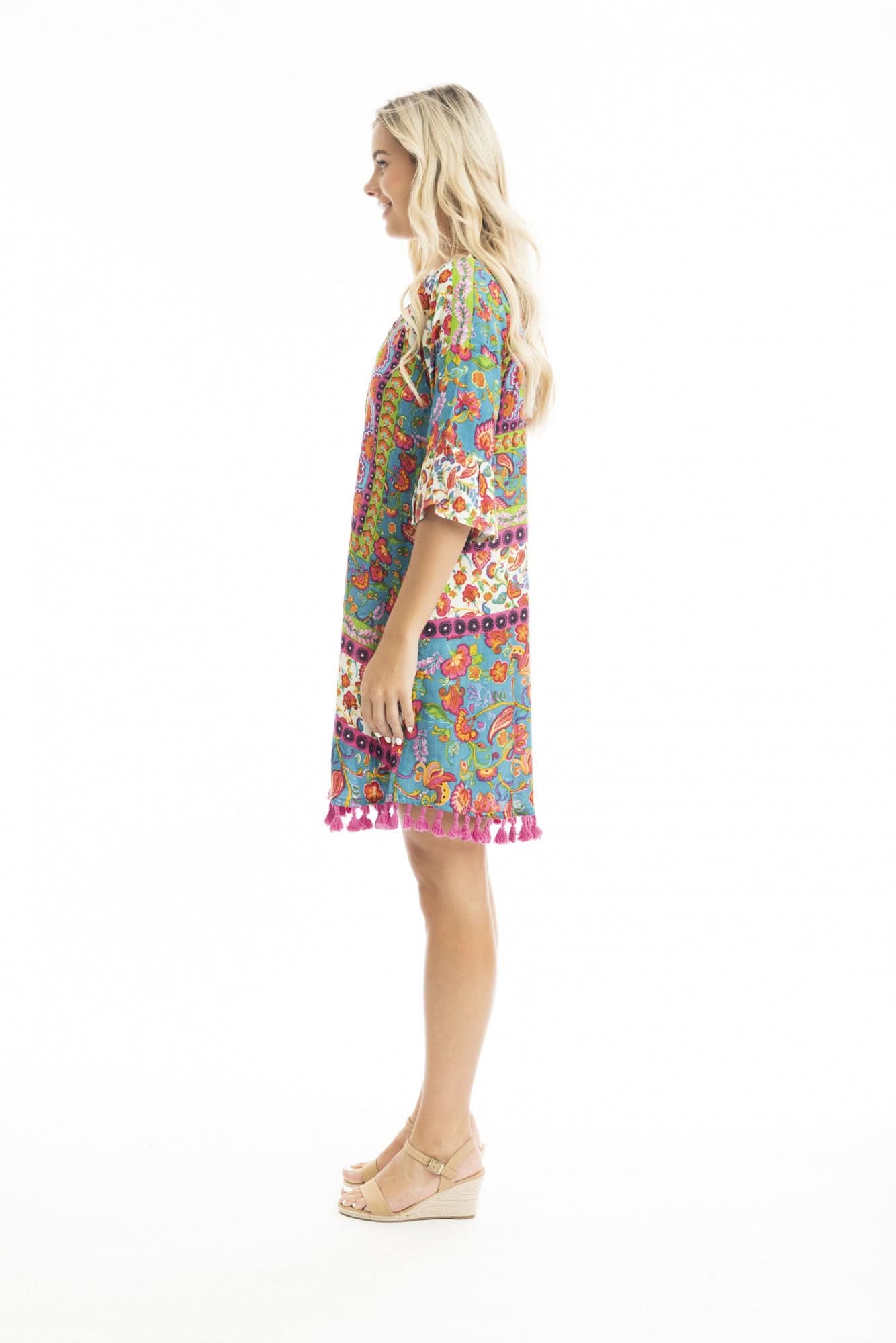 A woman in the vibrant Orientique Carvalho Tassel Dress with bell sleeves and pink tassel ties, paired with beige sandals, stands against a white background—an ideal summer look for warm weather occasions.