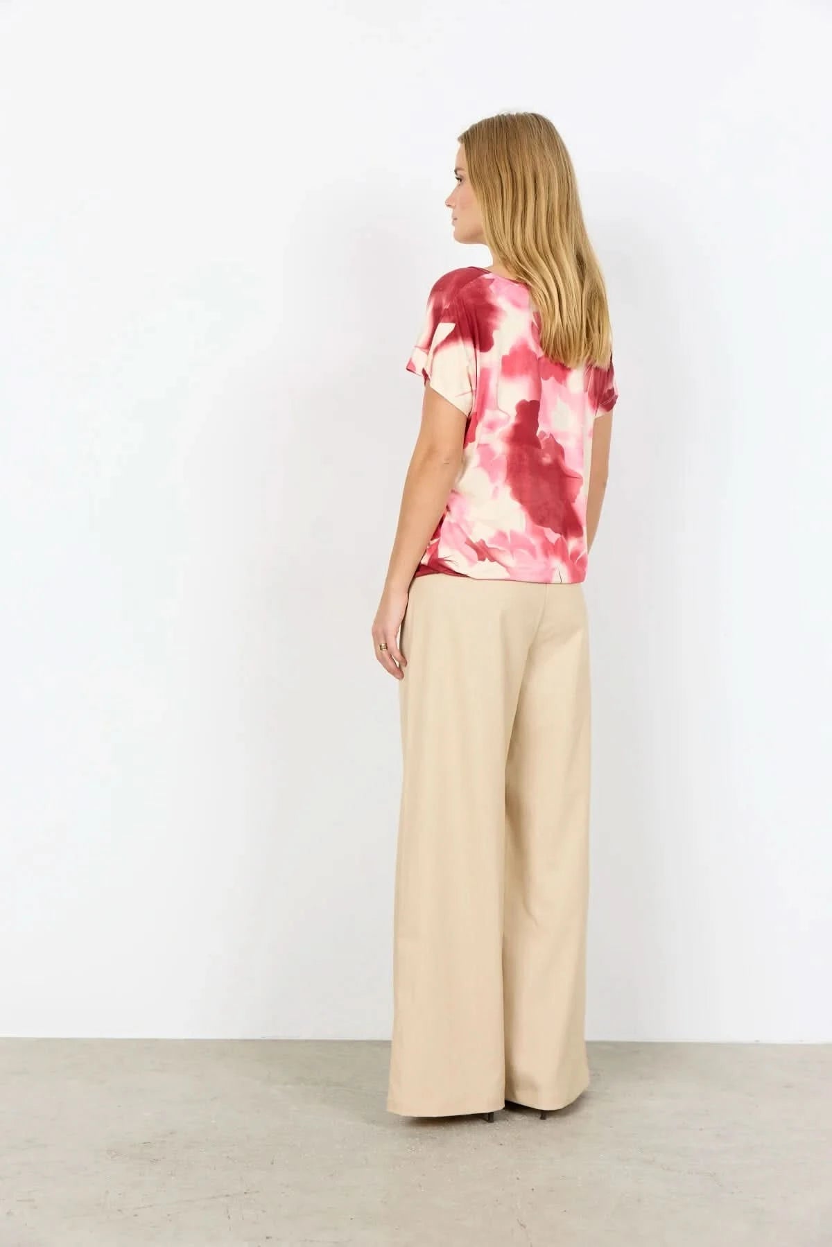 A woman faces away, wearing the pink and white tie-dye Marica Top by Soya Concepts with beige wide-leg pants, set against a plain white background.