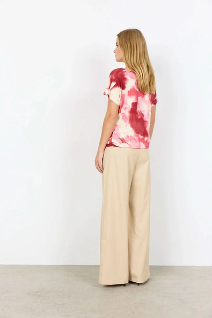 A woman faces away, wearing the pink and white tie-dye Marica Top by Soya Concepts with beige wide-leg pants, set against a plain white background.