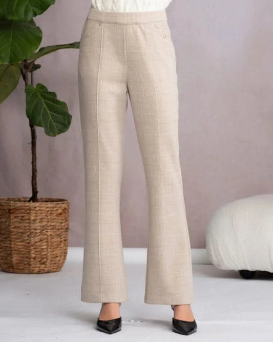 A person wearing Alison Sheri’s Front Seam Check Trouser, high-waisted and beige, paired with black pointed shoes, stands indoors by a plant and white cushioned seat—an elegant display of polished professional style.