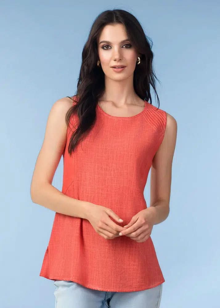 A woman with long dark hair wears Alison Sheri’s Easy Flow Sleeveless Top in coral with a V-neck and light blue jeans, standing against a plain light blue background.