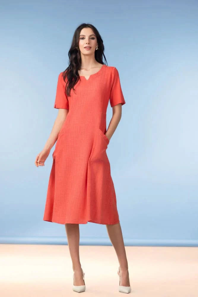 A woman wearing the Alison Sheri Margo Linen Blend Midi Dress, a coral A-line summer style with pockets, stands against a light blue background.