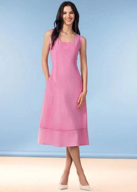 A woman stands against a light blue background wearing the Alison Sheri Maris Linen Blend Dress—a sleeveless, pink midi dress with a V-neckline—paired with white heels for a classic, timeless look.