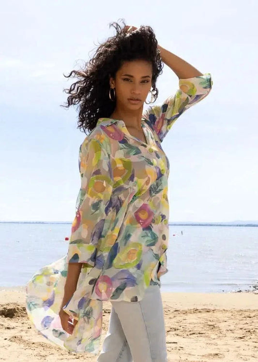 A woman wears the Alison Sheri Flowy Blouse with a colorful floral pattern and three-quarter sleeves, paired with light pants, while standing on a sandy beach with water and sky in the background.