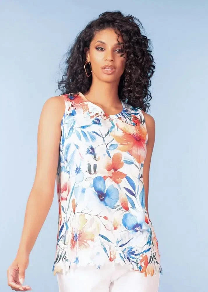 Woman with curly hair wearing the Alison Sheri Floral Lace Top—a sleeveless, floral print blouse in blue, orange, and white—standing against a light blue background.