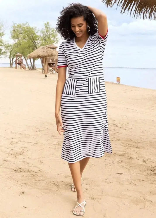 A woman strolls barefoot along a sandy beach, with trees and thatched huts behind her, wearing the Alison Sheri Navy/White Stripe Dress.