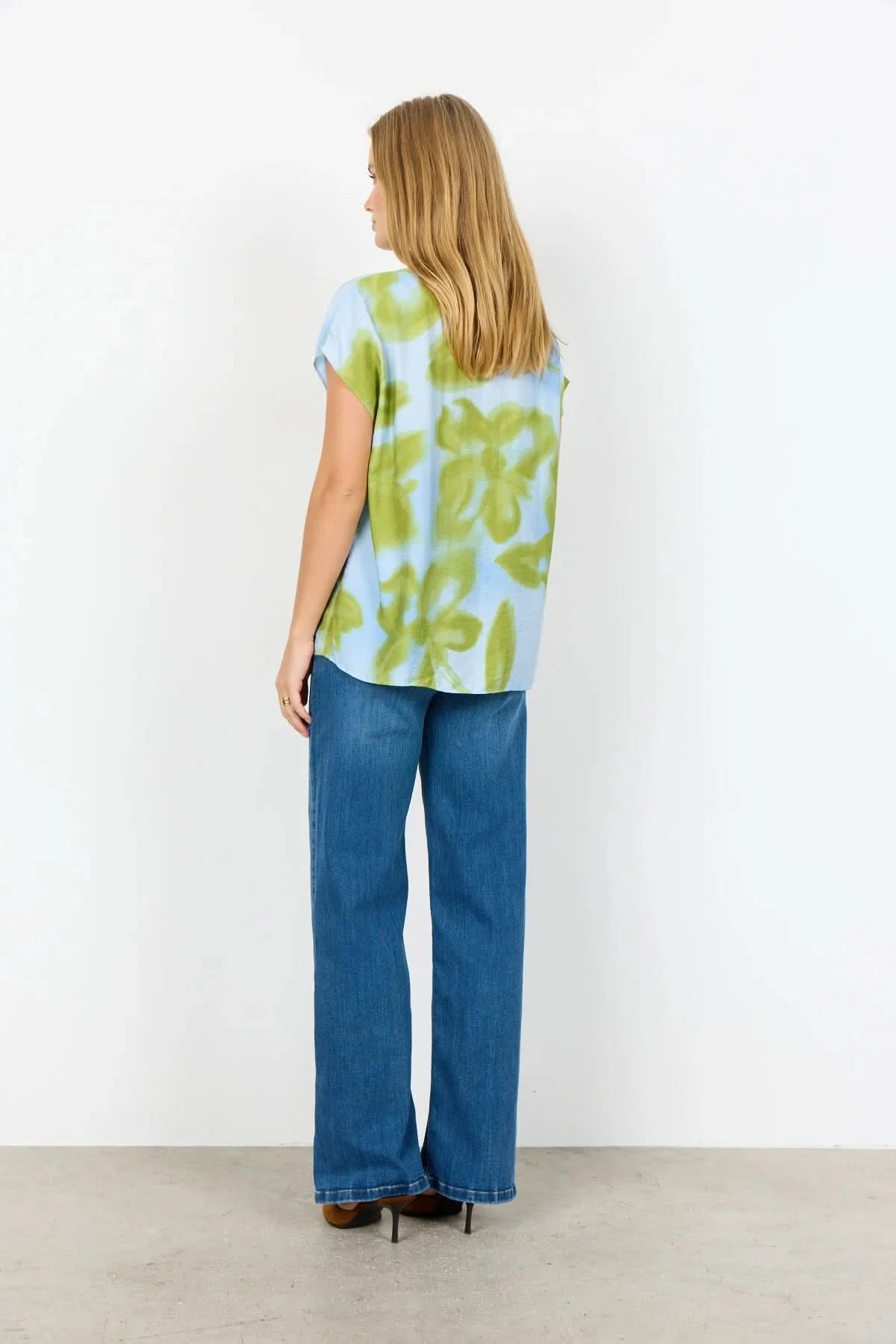 A woman stands against a white background wearing the Soya Concepts Cate Top, a sustainable blue and green floral print short-sleeve blouse, paired with blue jeans.