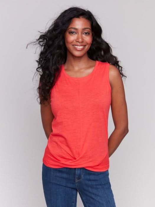 A woman with long, wavy dark hair wears the Charlie B Cotton Slub Tank Top With Knot Detail in red and blue jeans, standing against a light gray background while touching her hair.