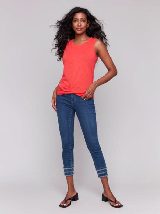 A woman with long, wavy dark hair wears the Charlie B Cotton Slub Tank Top With Knot Detail in red and blue jeans, standing against a light gray background while touching her hair.