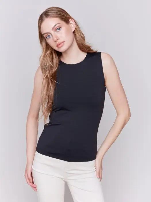 A person with long curly hair, wearing a Charlie B Super Stretch Sleeveless Top, blue jeans, and black ankle boots, stands posing against a plain background.